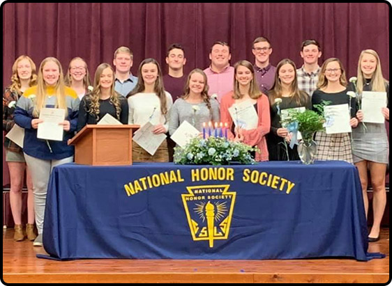 Fifteen students stand behind a table with a blue tablecloth that says National Honor Society. The students are holding certificates.